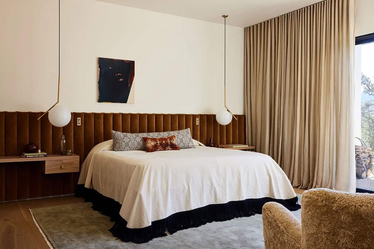 Primary bedroom with full-wall channel-tufted tobacco velvet headboard, brass globe pendants, cream bedding with fringe hem, and mountain views