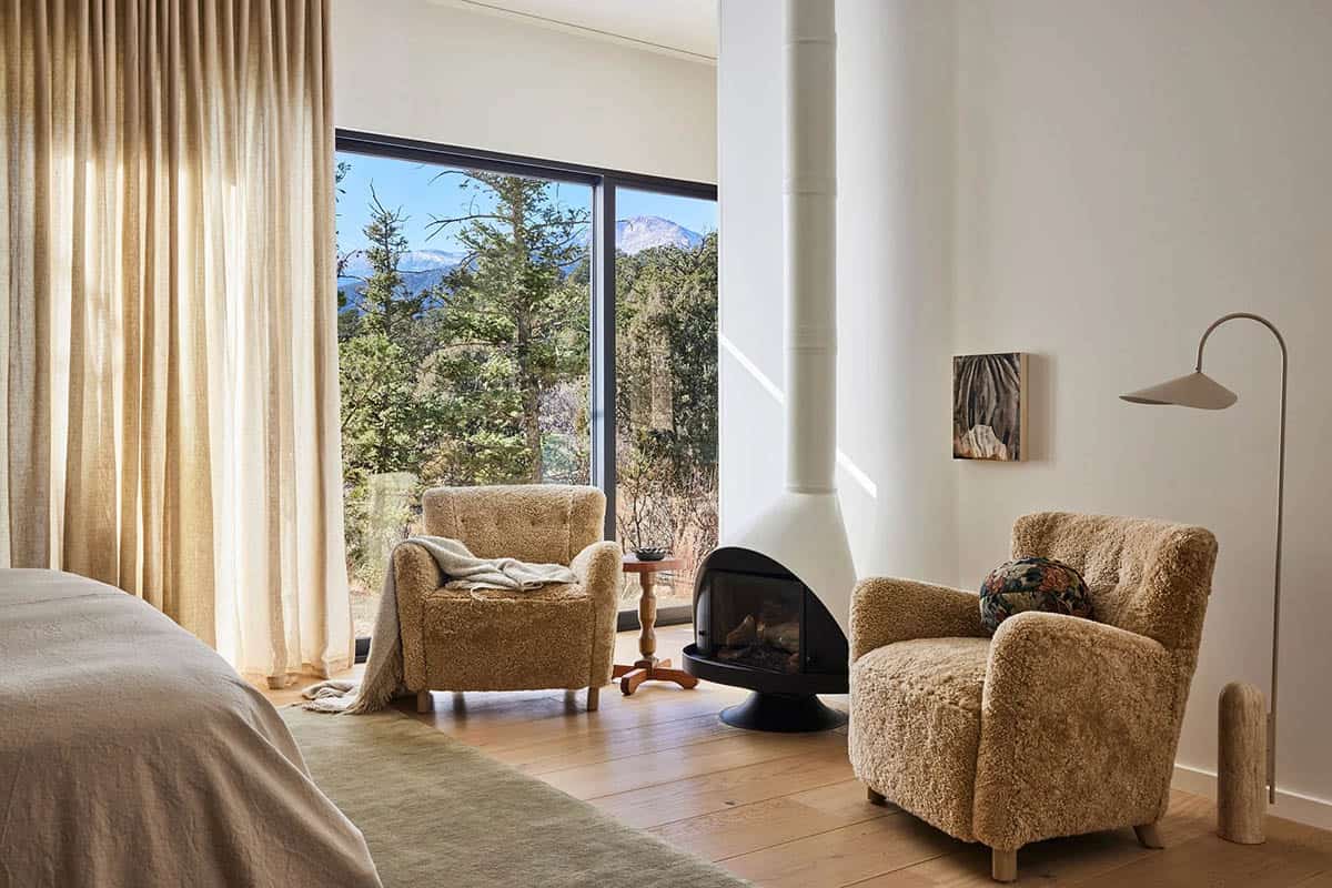 Primary bedroom sitting area with two shearling armchairs, freestanding black fireplace, floor-to-ceiling windows with mountain and forest views