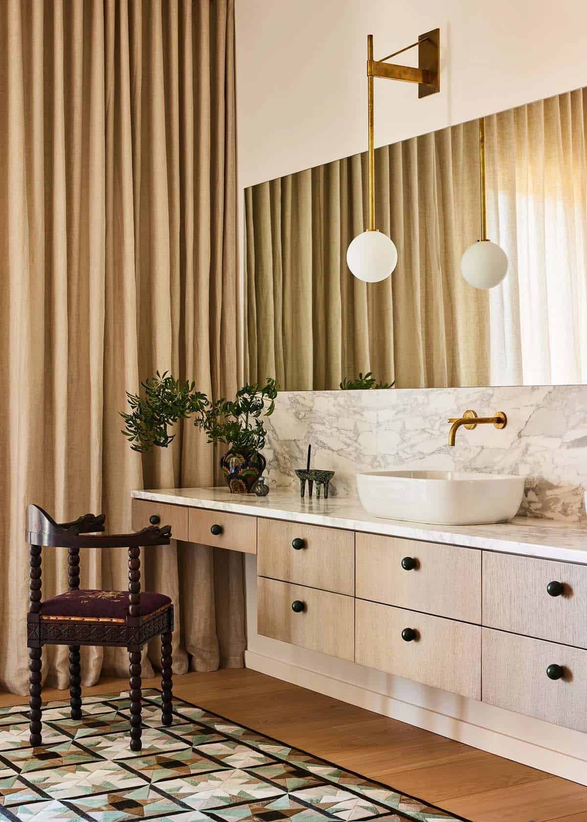 Primary bathroom vanity with floating oak cabinet, marble countertop, vessel sink, brass globe sconce, and geometric tile floor beside an antique spool chair