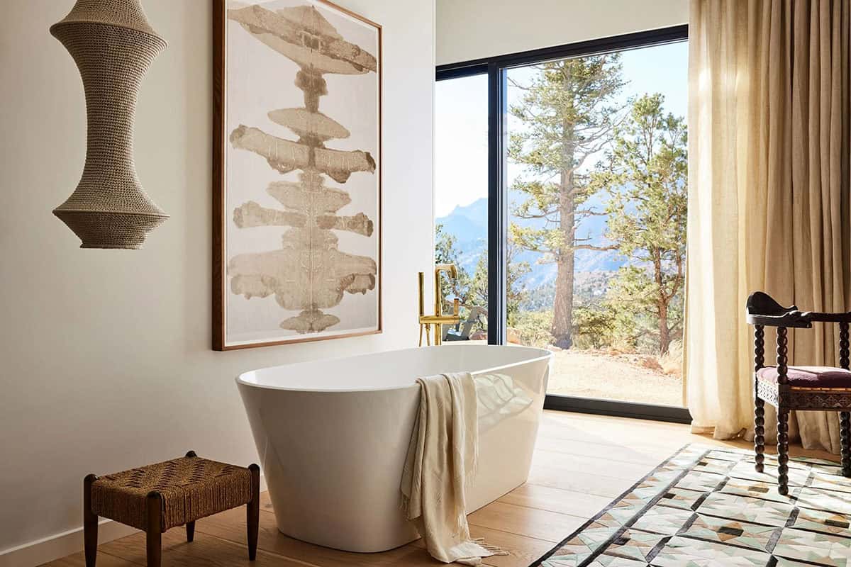 Primary bathroom with freestanding white soaking tub, brass floor-mount faucet, large abstract artwork, and floor-to-ceiling mountain view window