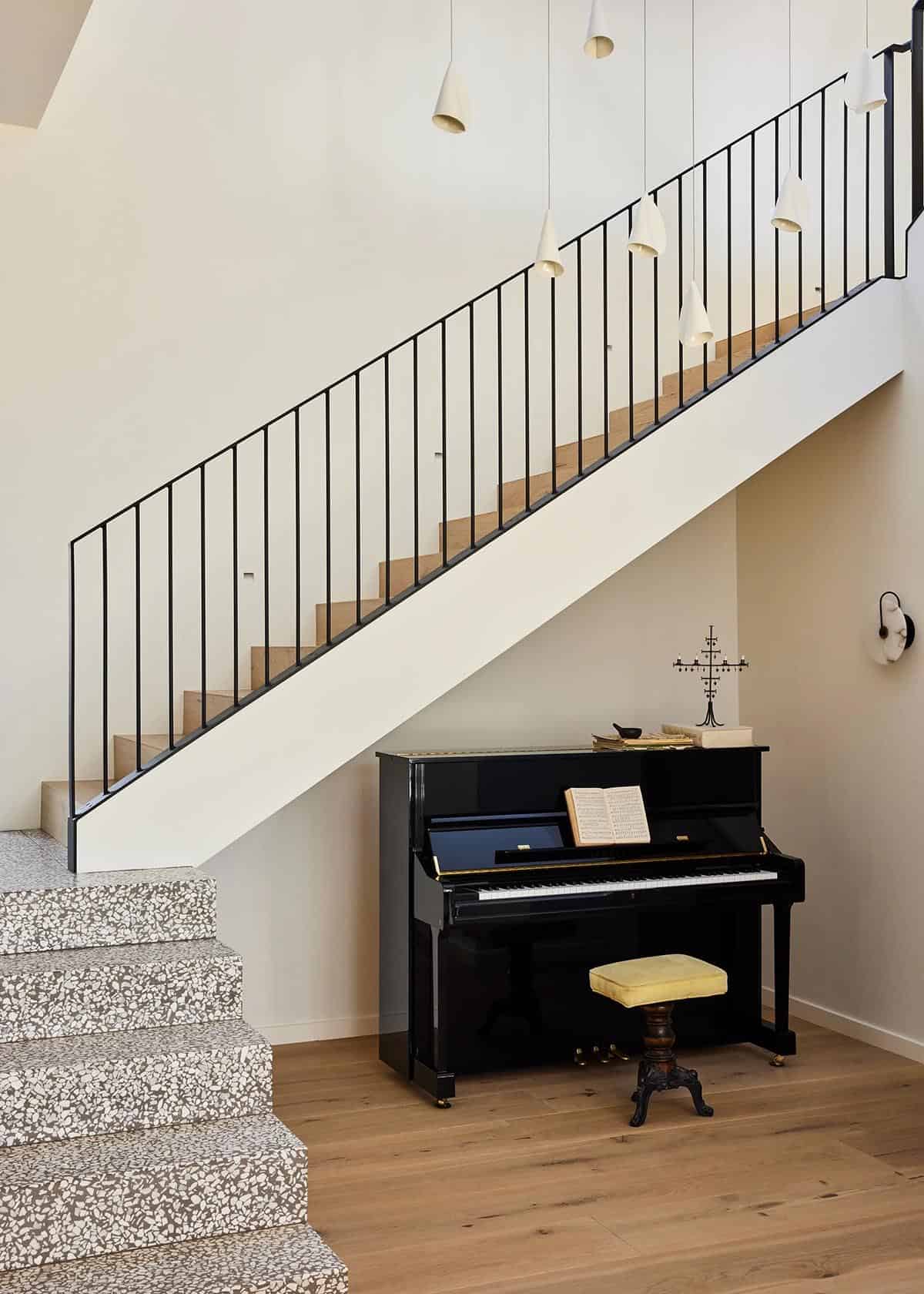 Open staircase with black steel balustrade, terrazzo lower steps, oak upper treads, and an upright piano tucked underneath with cone pendant lights