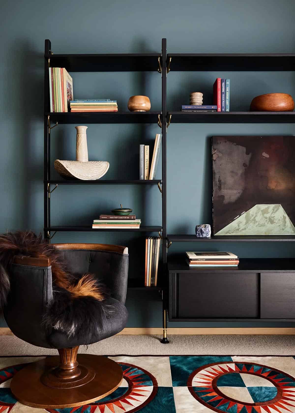den with deep teal-painted walls, black metal shelving unit with books and ceramics, colorful geometric rug, and a dark velvet swivel chair