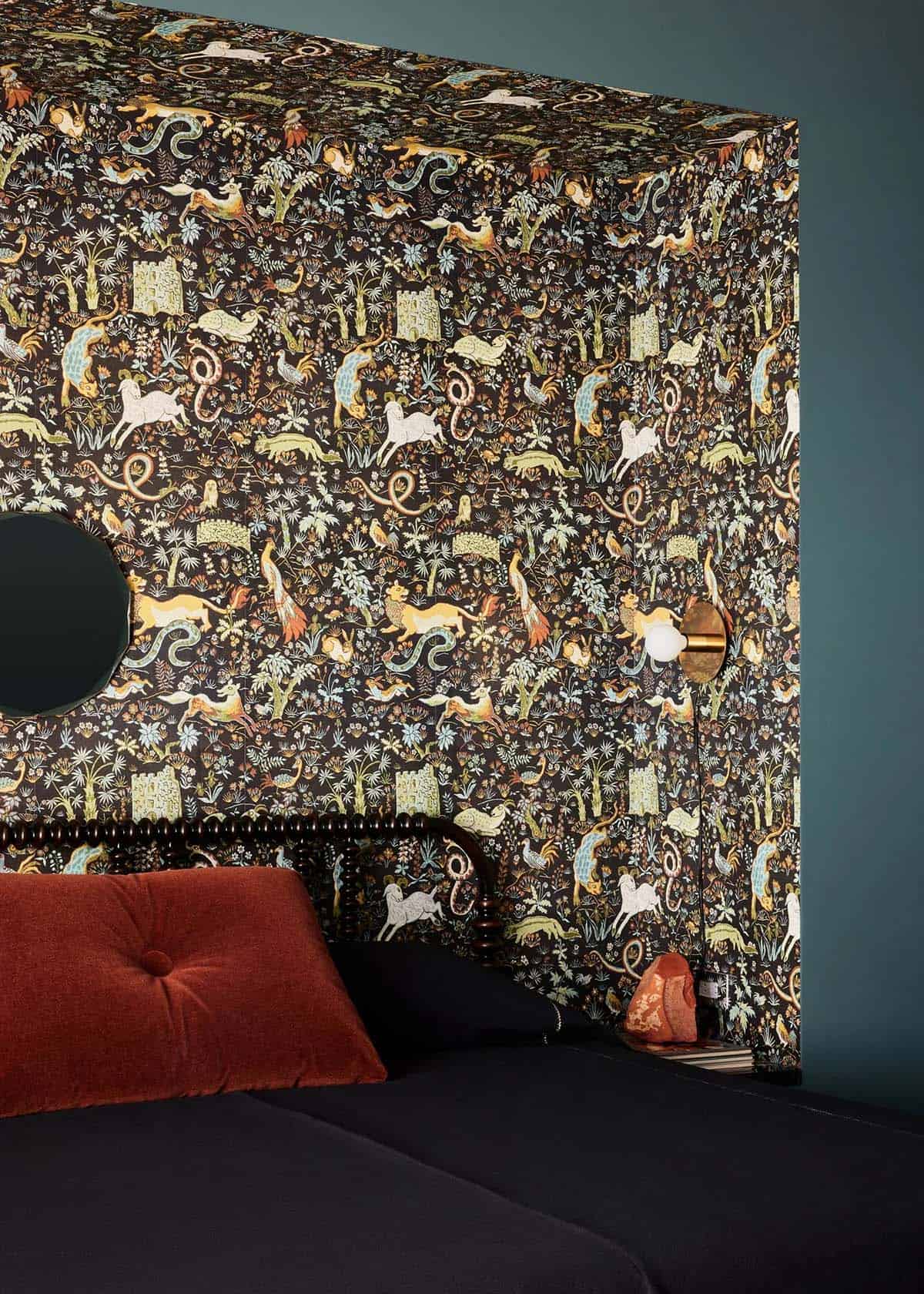 Close-up of floor-to-ceiling animal-print wallpaper lining a bed alcove with a brass globe sconce and rust-colored pillow
