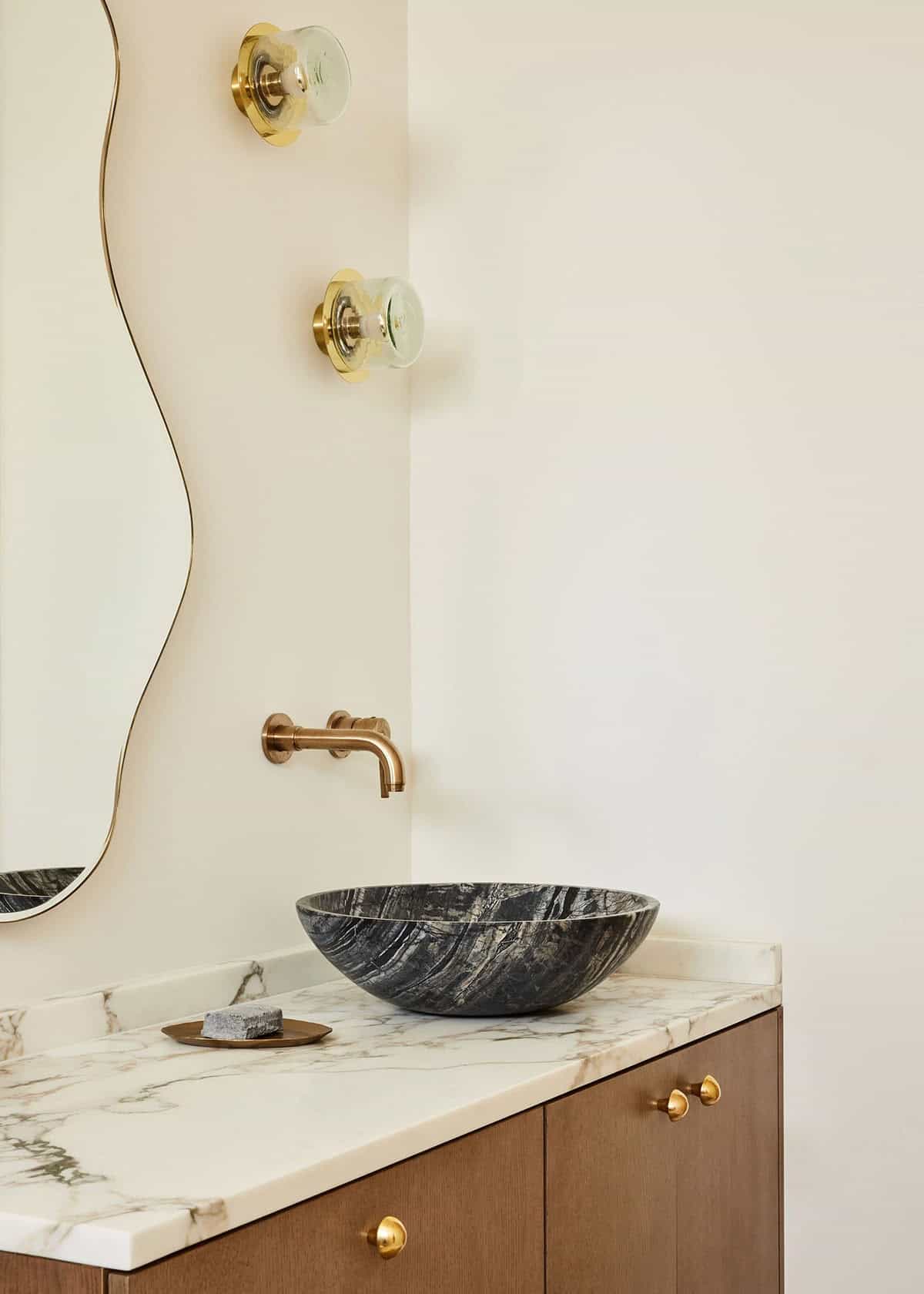 Bathroom vanity detail with dark veined marble vessel sink, brass wall-mount faucet, flower-shaped brass wall sconces, and organic mirror