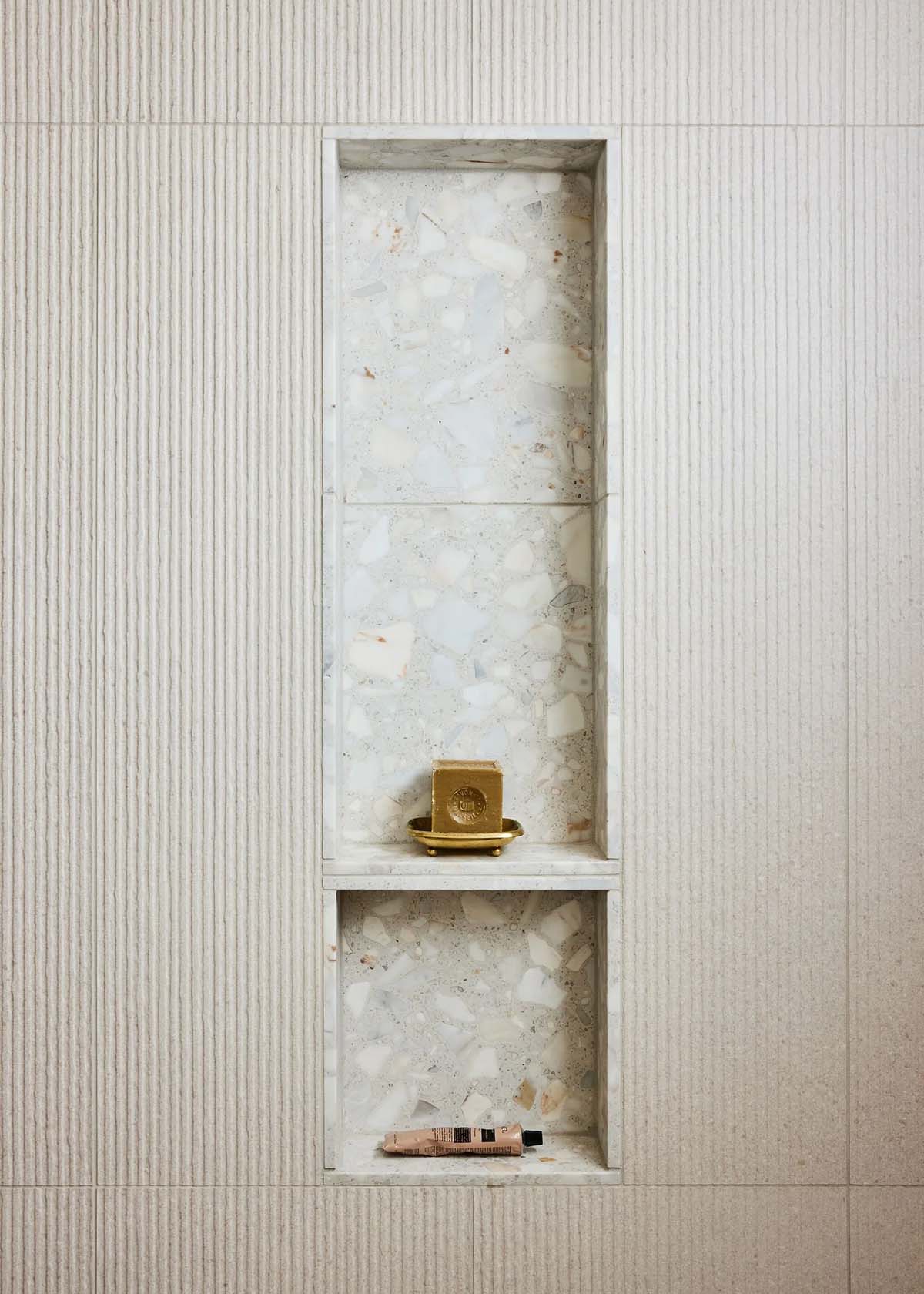 Shower niche detail lined with large-chunk terrazzo tile, holding a brass soap dish and toiletries, set into ribbed limestone wall tile