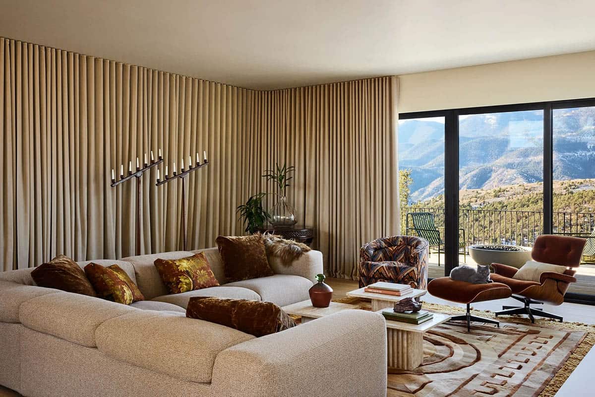 Upper-level lounge with cream sectional sofa, rust velvet pillows, travertine coffee table, Eames lounge chair, and dramatic mountain views through floor-to-ceiling glass doors