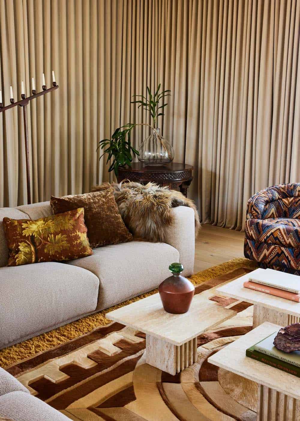 Lounge room detail showing cream sofa with jewel-toned pillows, travertine coffee tables, patterned armchair, and floor-length draped curtains