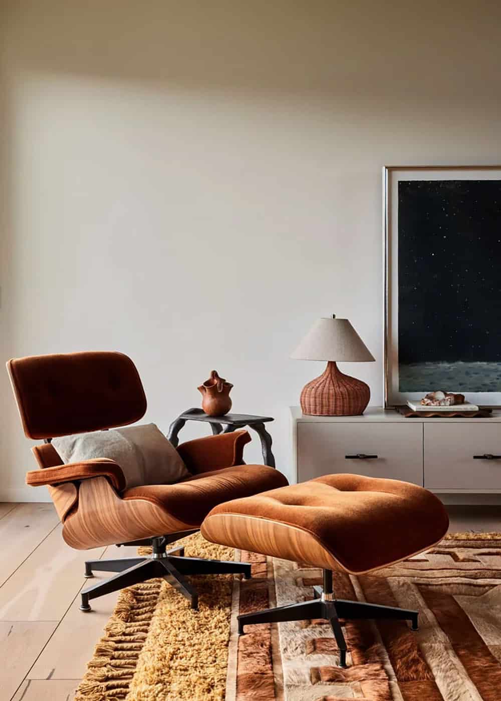 Rust velvet Eames lounge chair and ottoman on a textured rug beside a white credenza with a woven lamp and dark night-sky painting