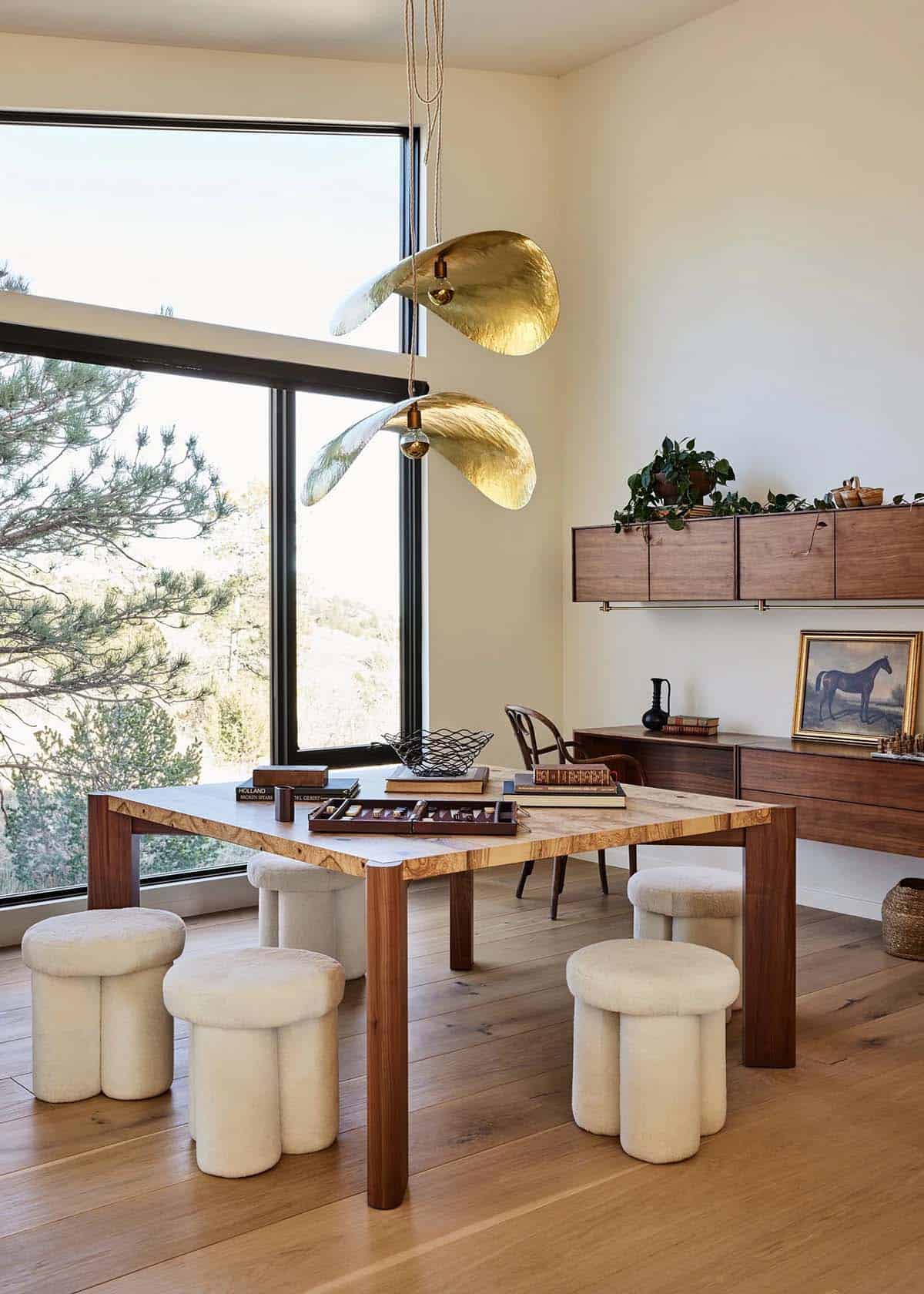Games room with live-edge wood table, shearling stools, sculptural brass leaf pendant light, and walnut wall-mounted shelving unit