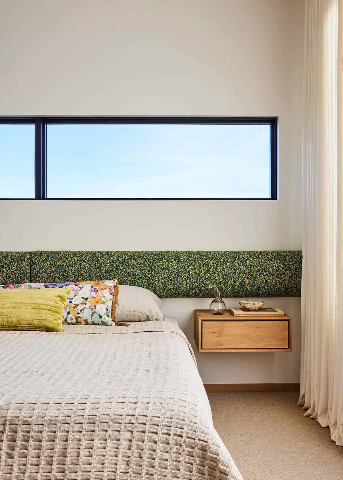 Guest bedroom headboard detail with green floral fabric, floating oak nightstand, horizontal clerestory window with blue sky view