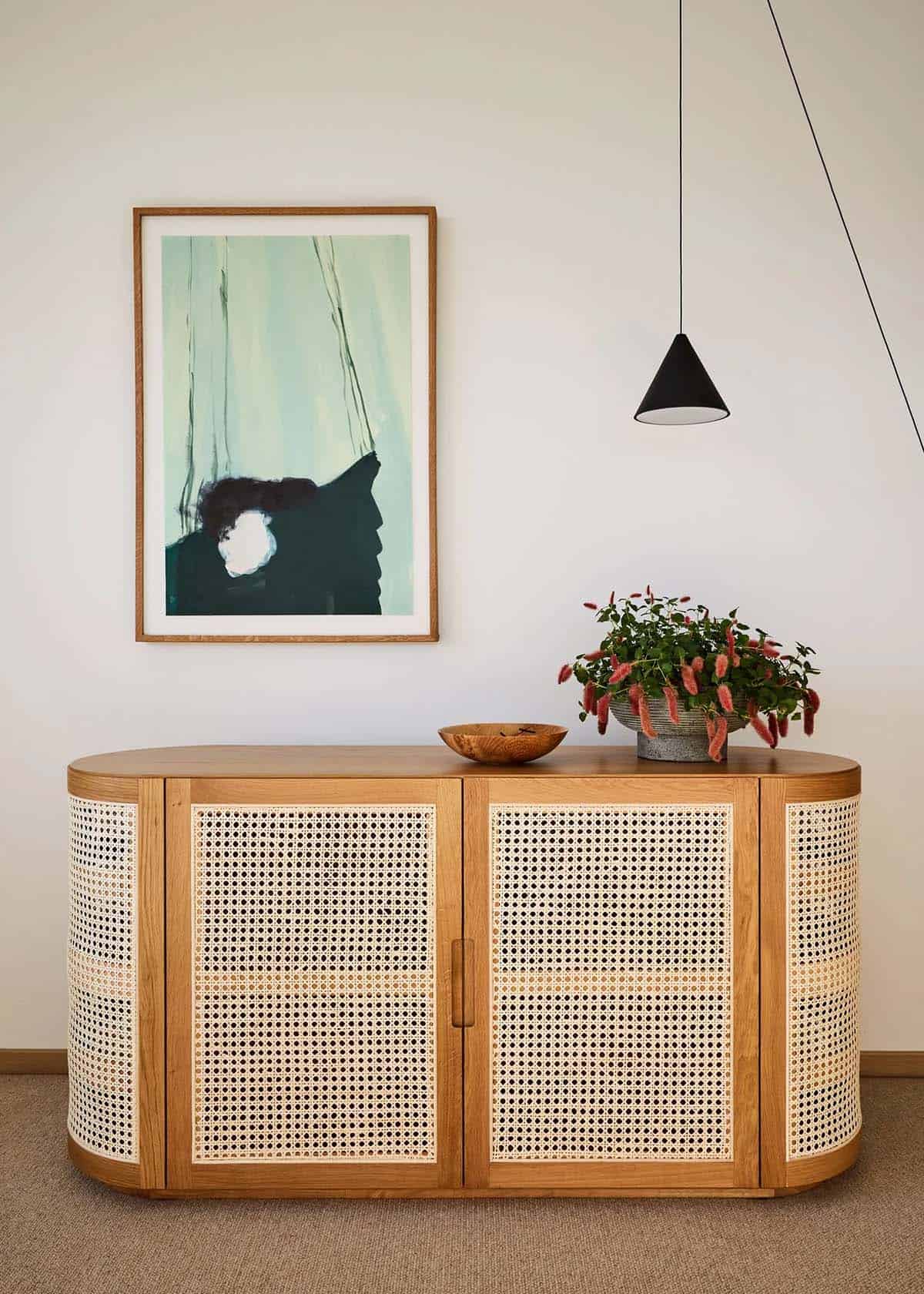 Guest bedroom corner with rounded oak and cane sideboard, wood bowl, potted flowering plant, abstract green artwork, and black cone pendant light