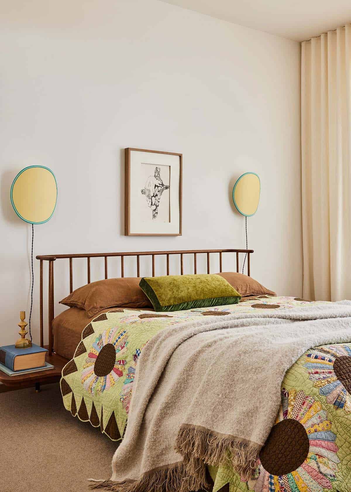 Bedroom with dark wood spindle bed frame, vintage daisy quilt, green velvet pillow, round yellow wall sconces, and framed figure drawing