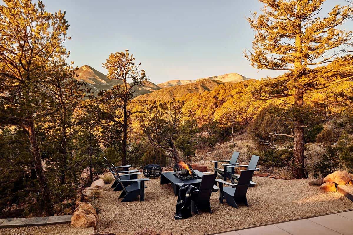 Outdoor fire pit patio with black modern Adirondack chairs surrounded by pine trees glowing in warm golden sunset light and Colorado mountain range beyond
