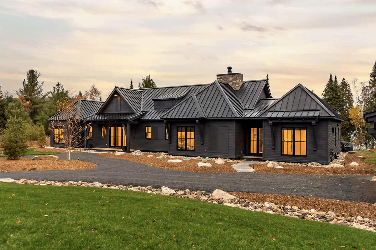 Street-facing view of dark modern cottage with glowing windows and stone chimney at dusk