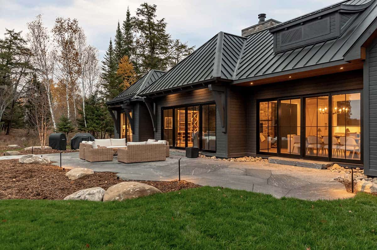 Rear patio of dark cottage with wicker furniture, large windows revealing lit interior at dusk
