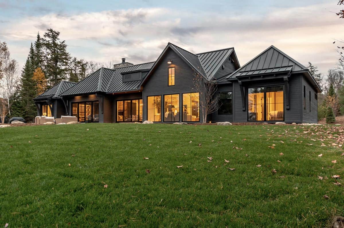 Lakeside rear facade of black modern farmhouse glowing at twilight with expansive lawn