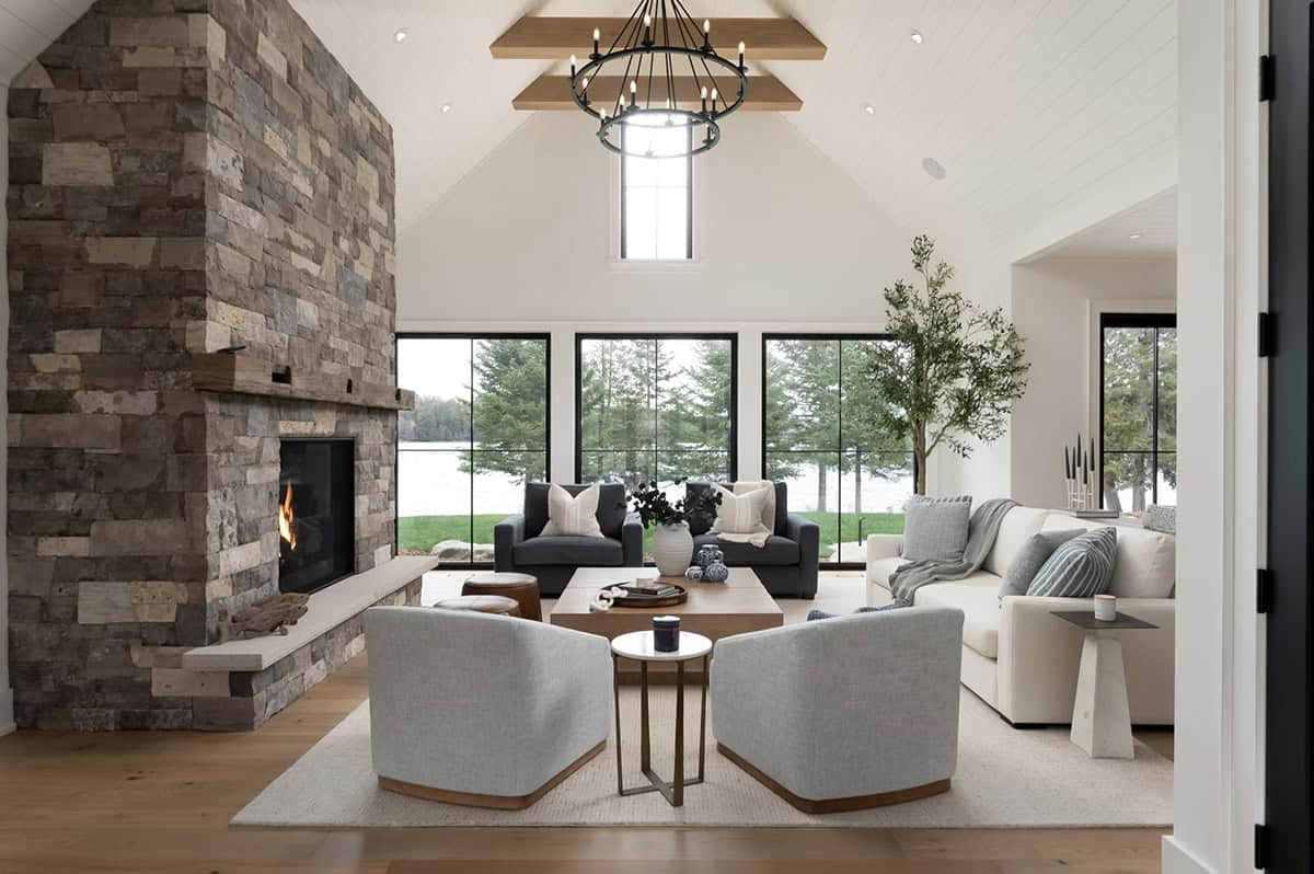 Vaulted great room with floor-to-ceiling stone fireplace, wood beams, and lake views through black-framed windows