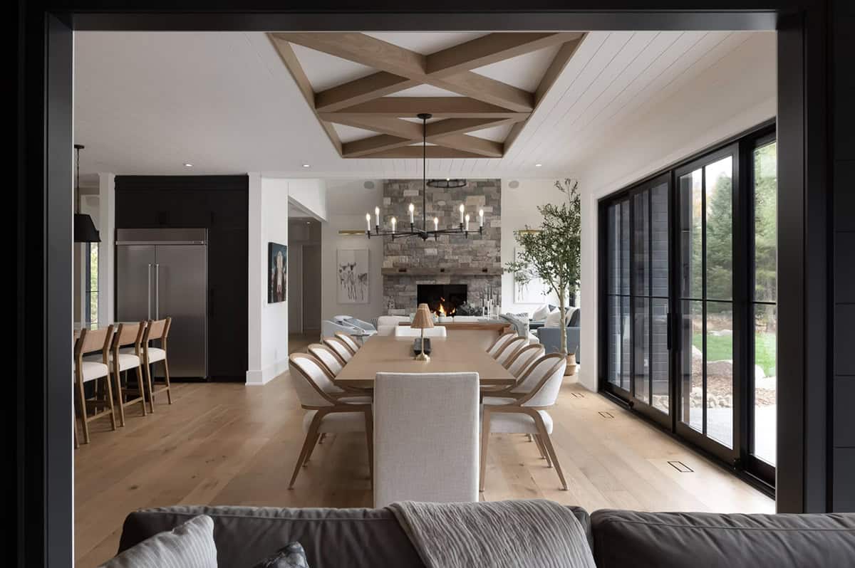 Dining room framed by dark doorway with geometric beam ceiling, long table, stone fireplace, and kitchen beyond