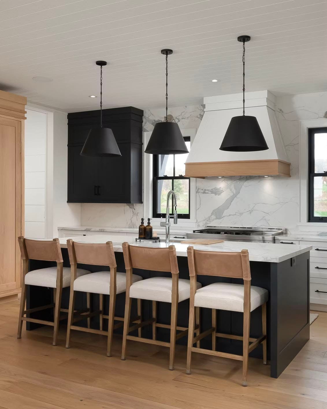 Kitchen island with leather barstools, marble backsplash, wood-accented range hood, and black pendant lights