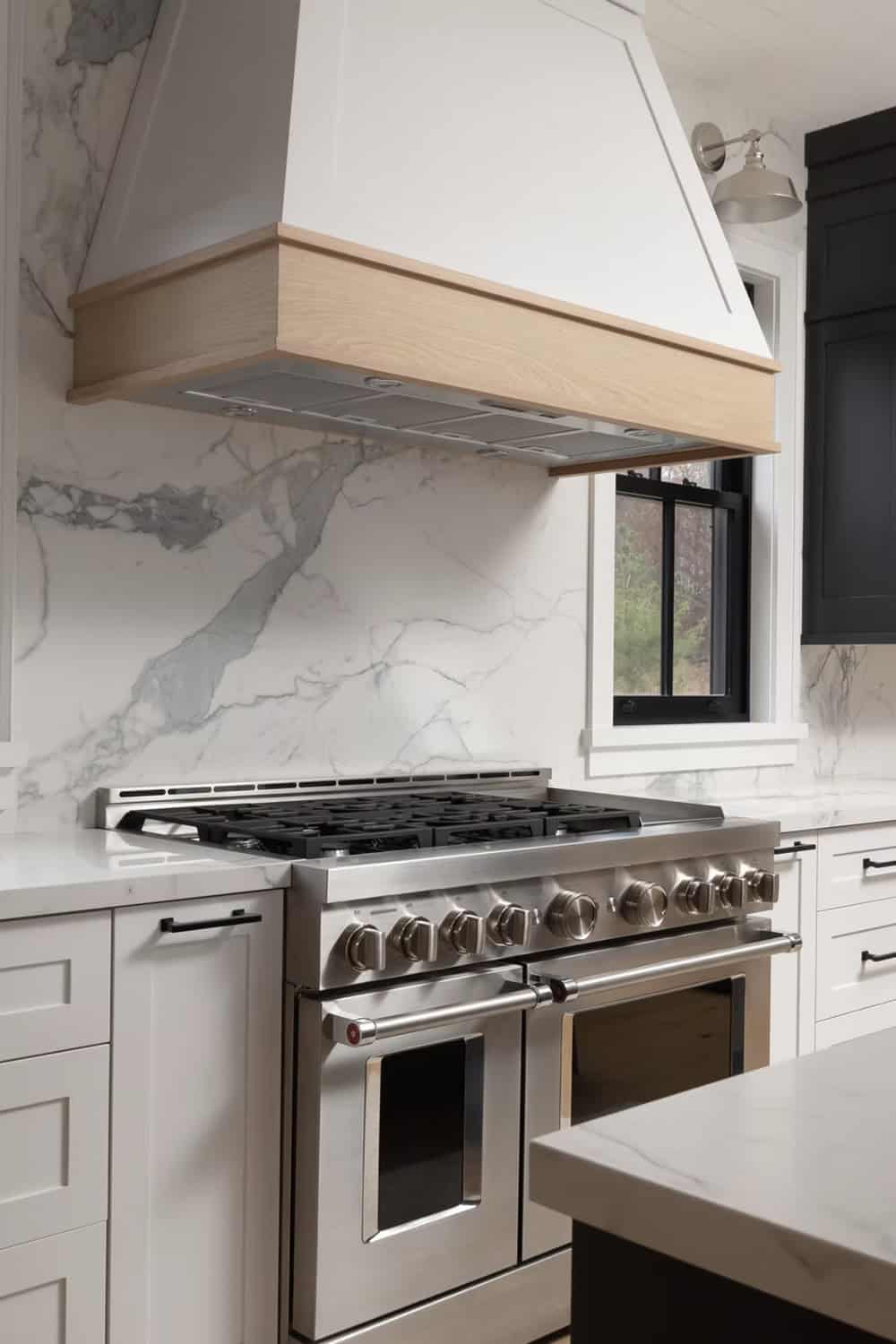 Close-up of stainless gas range with wood-trimmed white range hood and full-height marble slab backsplash