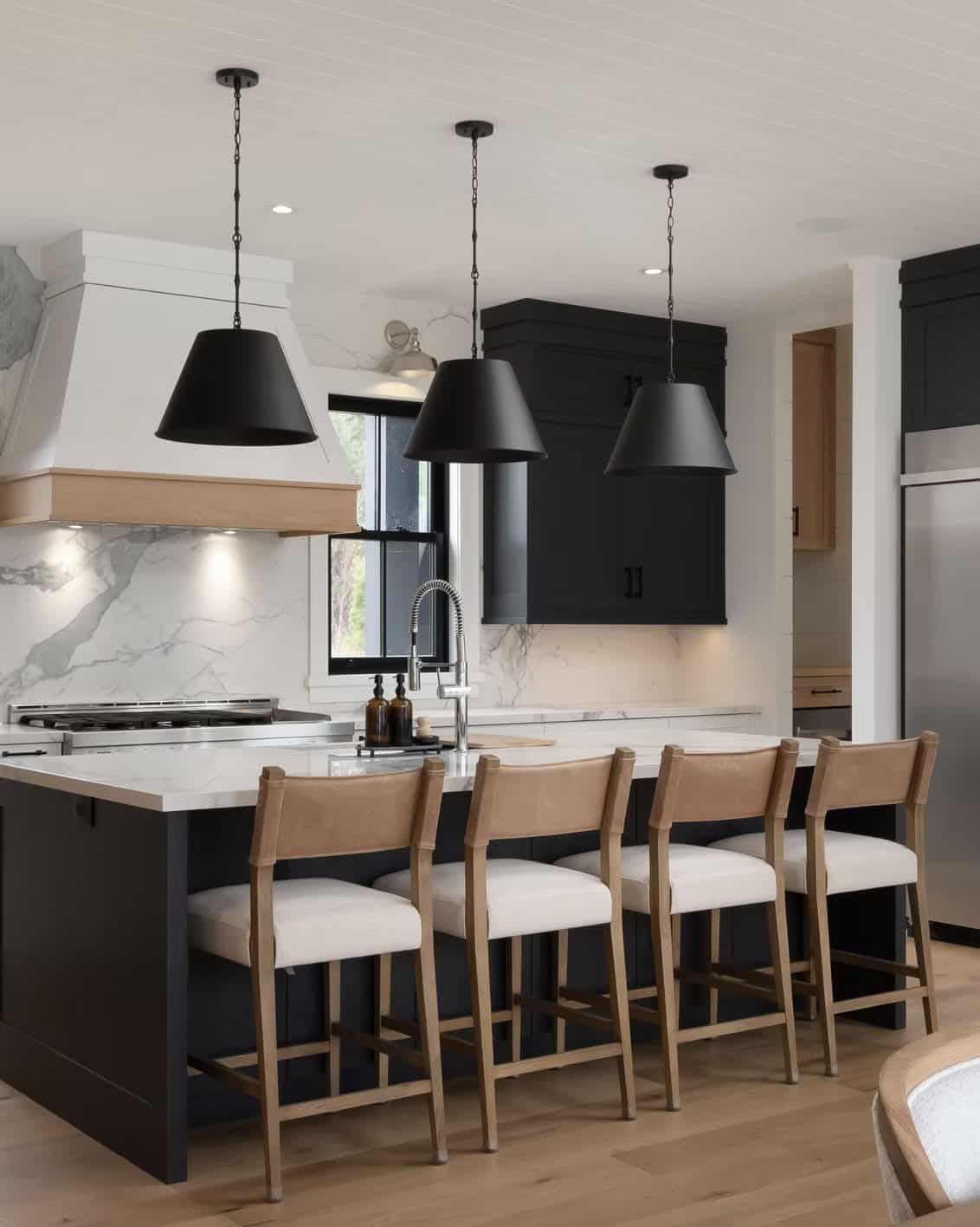Dark kitchen island with leather barstools, white range hood, marble backsplash, and black pendant trio