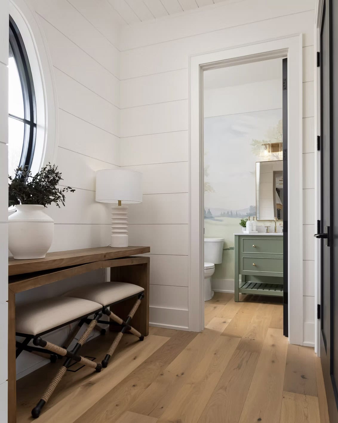 Bright cottage foyer nook with console table and oval window opening to powder room with green vanity