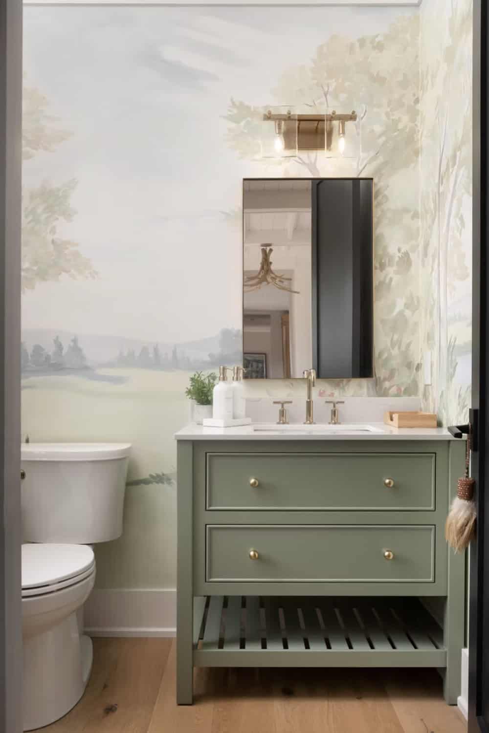 Powder room with sage green vanity, brass fittings, oval mirror, and watercolor landscape mural wallpaper
