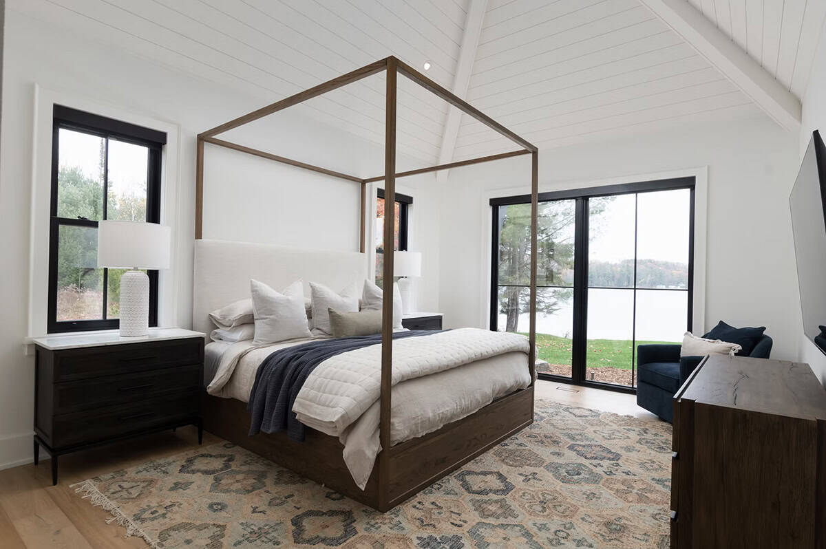 Primary bedroom with walnut four-poster canopy bed, lake view through black-framed glass doors, and patterned rug