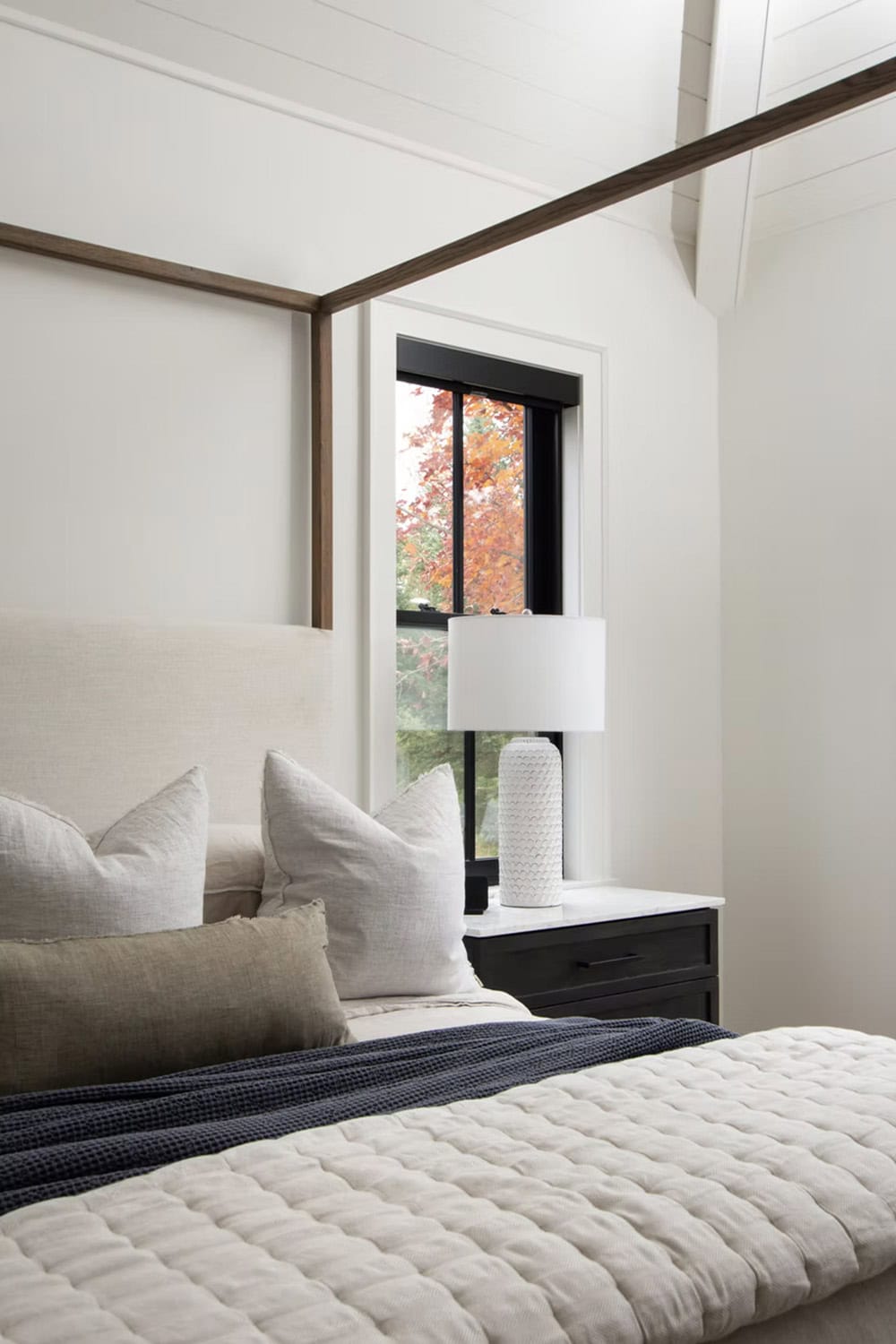 Close-up of canopy bed headboard with layered pillows, dark nightstand, and black-framed window with fall foliage