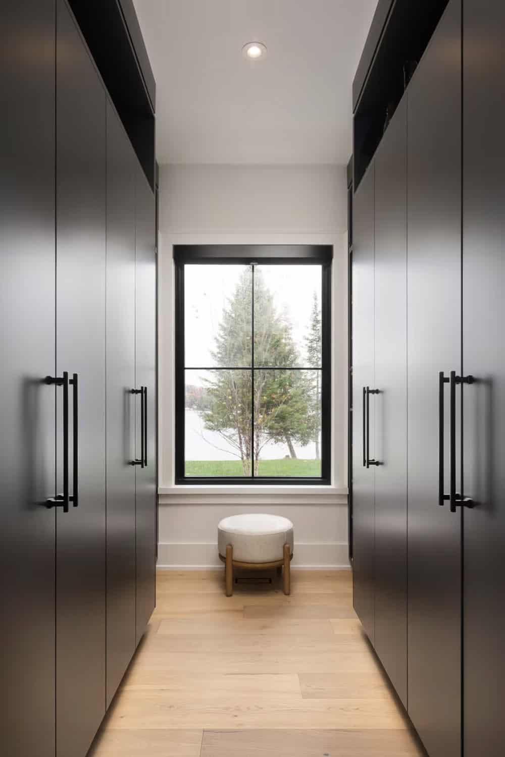Walk-in closet with dark floor-to-ceiling wardrobe cabinets, black hardware, and lake view window with ottoman