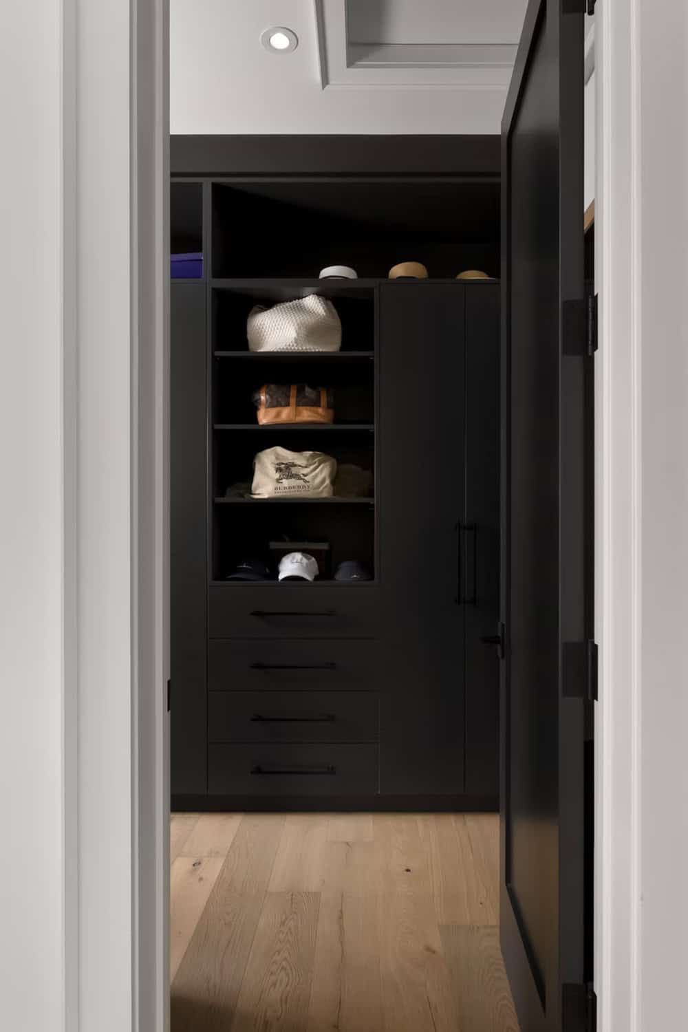 Dark walk-in closet with open shelving, bags, hats, and built-in drawers viewed through doorway