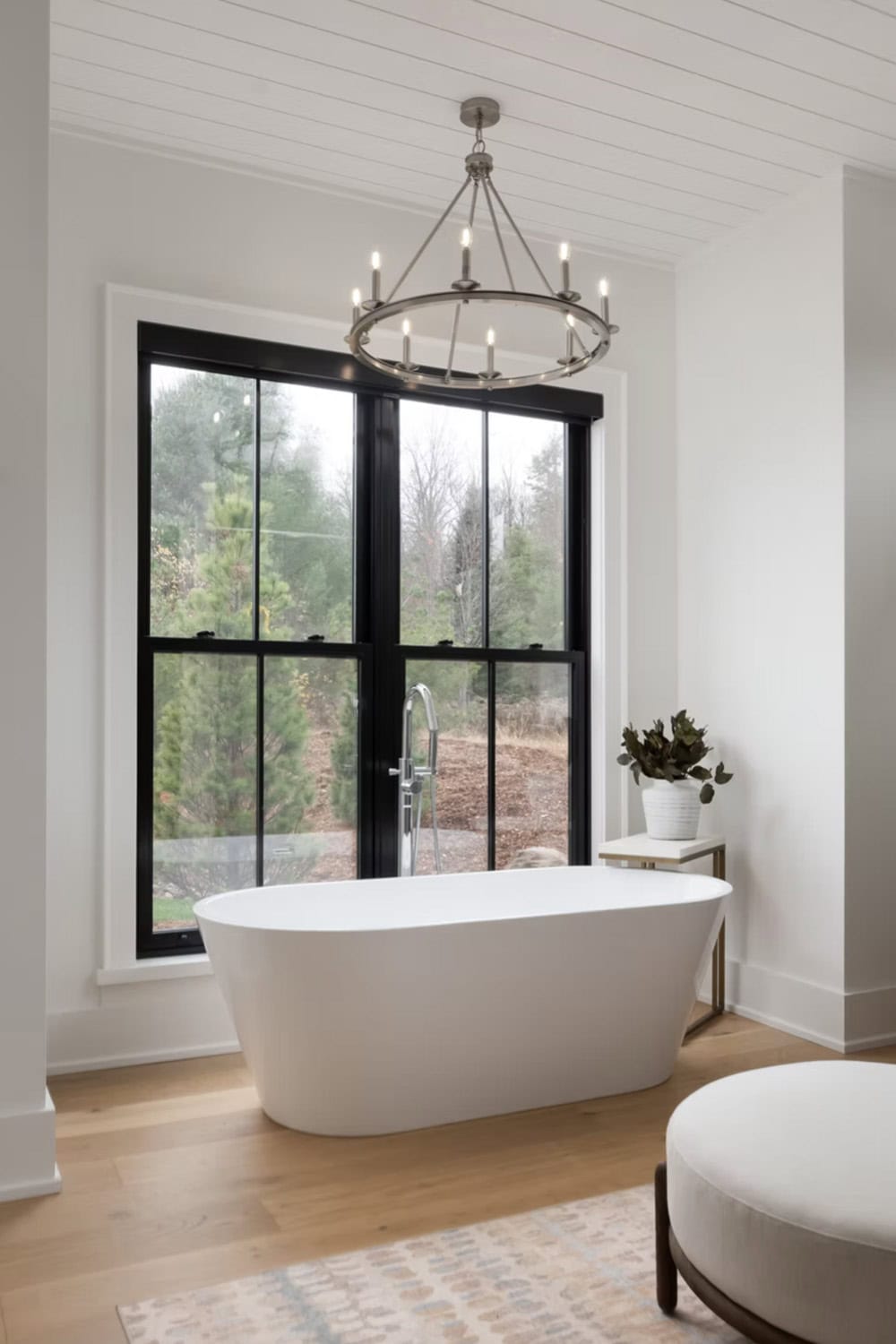 Freestanding white soaker tub beside large black-framed window with round chandelier and forest view