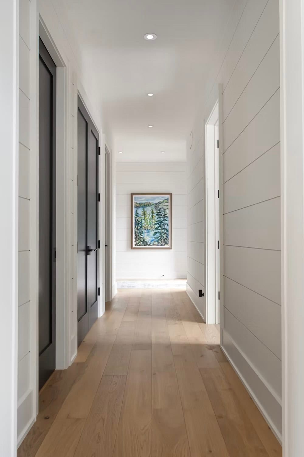 Bright shiplap hallway with black interior doors, oak floors, and landscape painting at the end