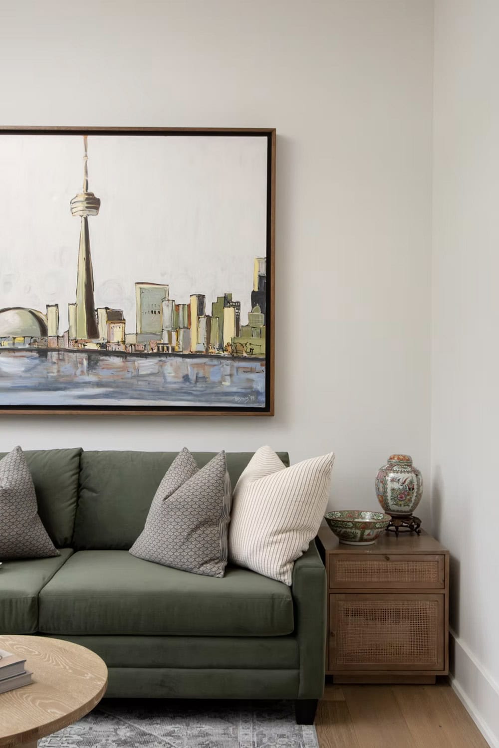 Close-up of green sofa with pillows beneath large Toronto skyline painting and rattan side cabinet