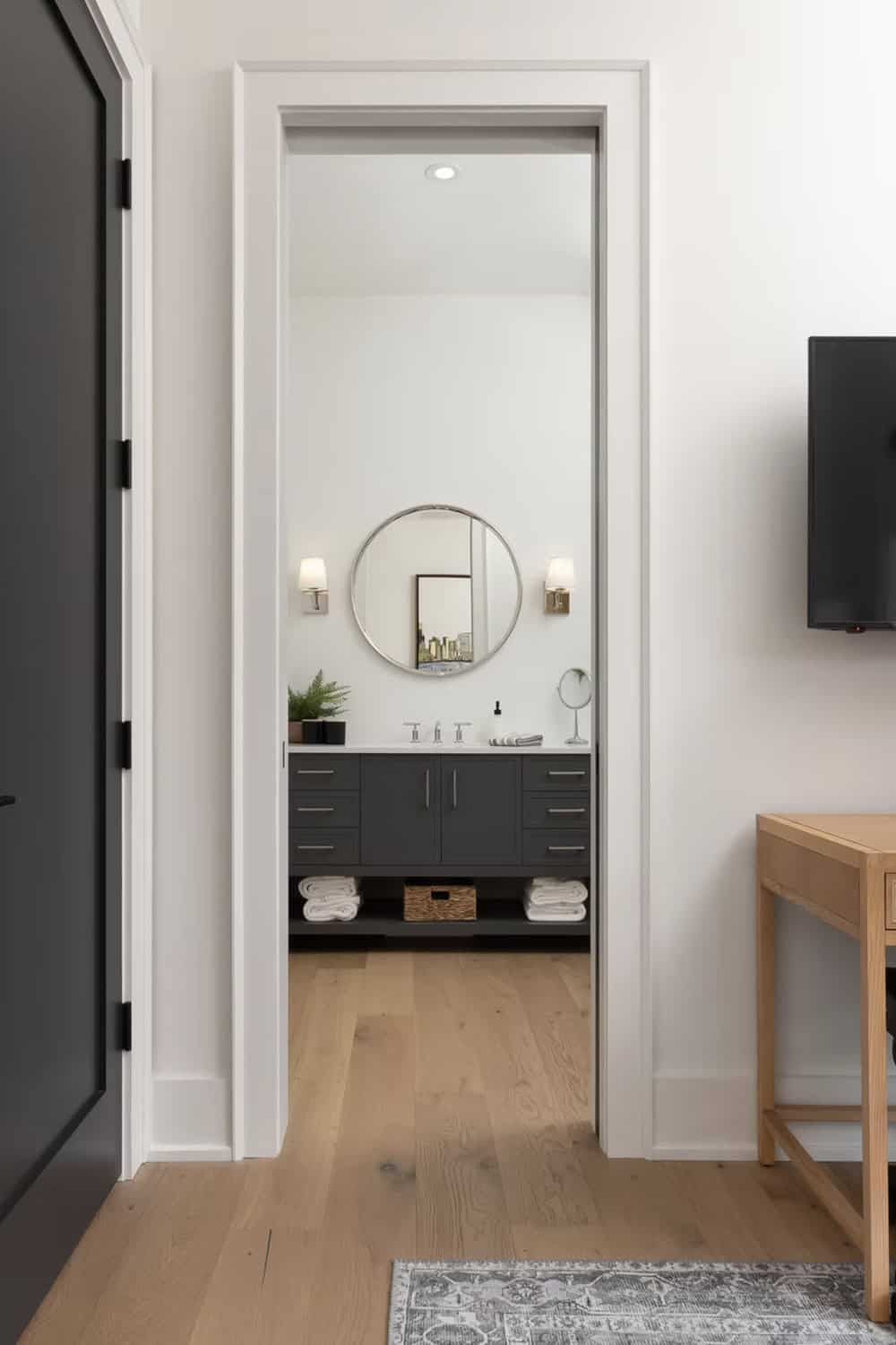 Bedroom doorway view into ensuite bathroom with dark vanity, round mirror, and wall sconces
