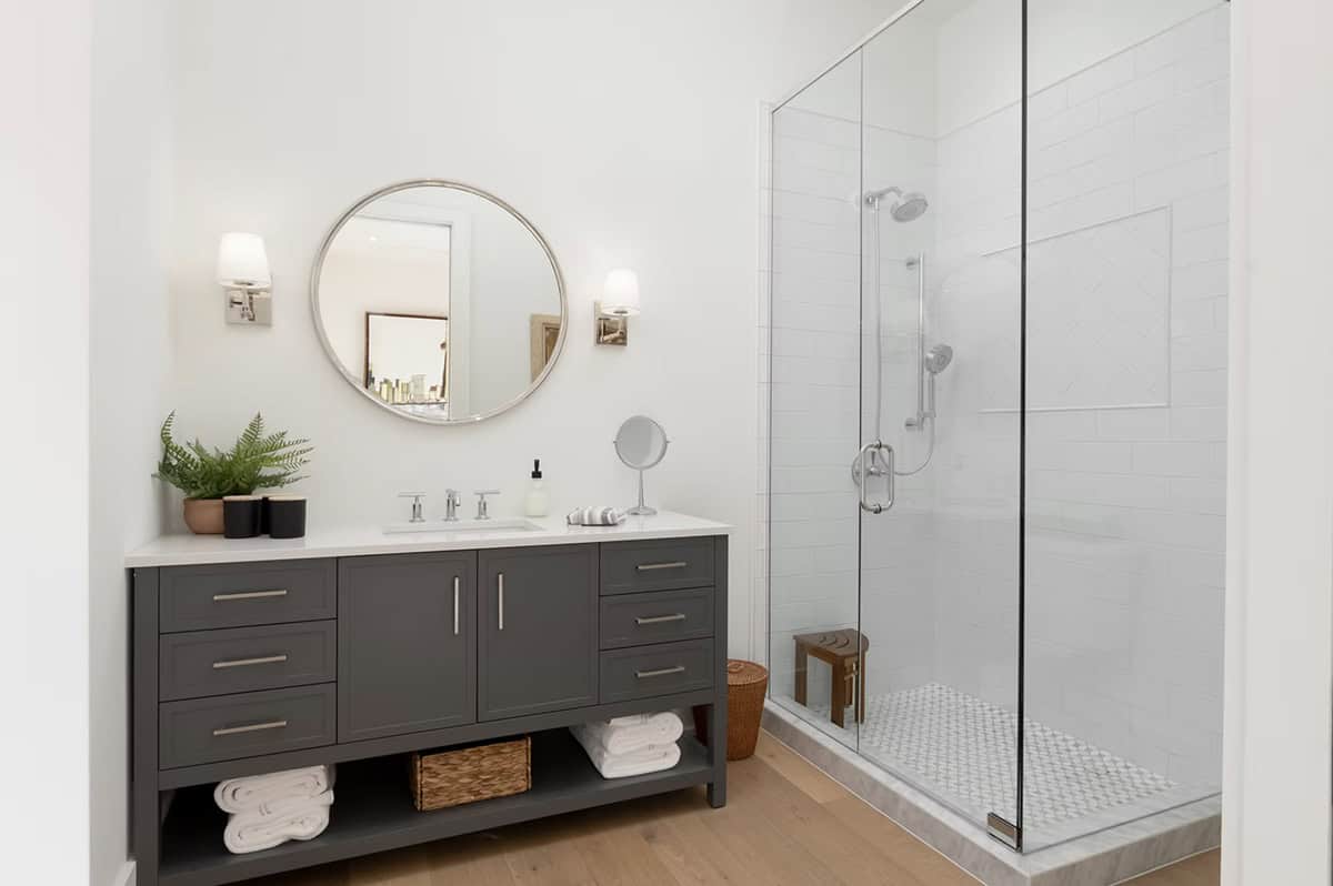 Guest bathroom with dark vanity, round mirror, wall sconces, and glass-enclosed shower with patterned floor tile