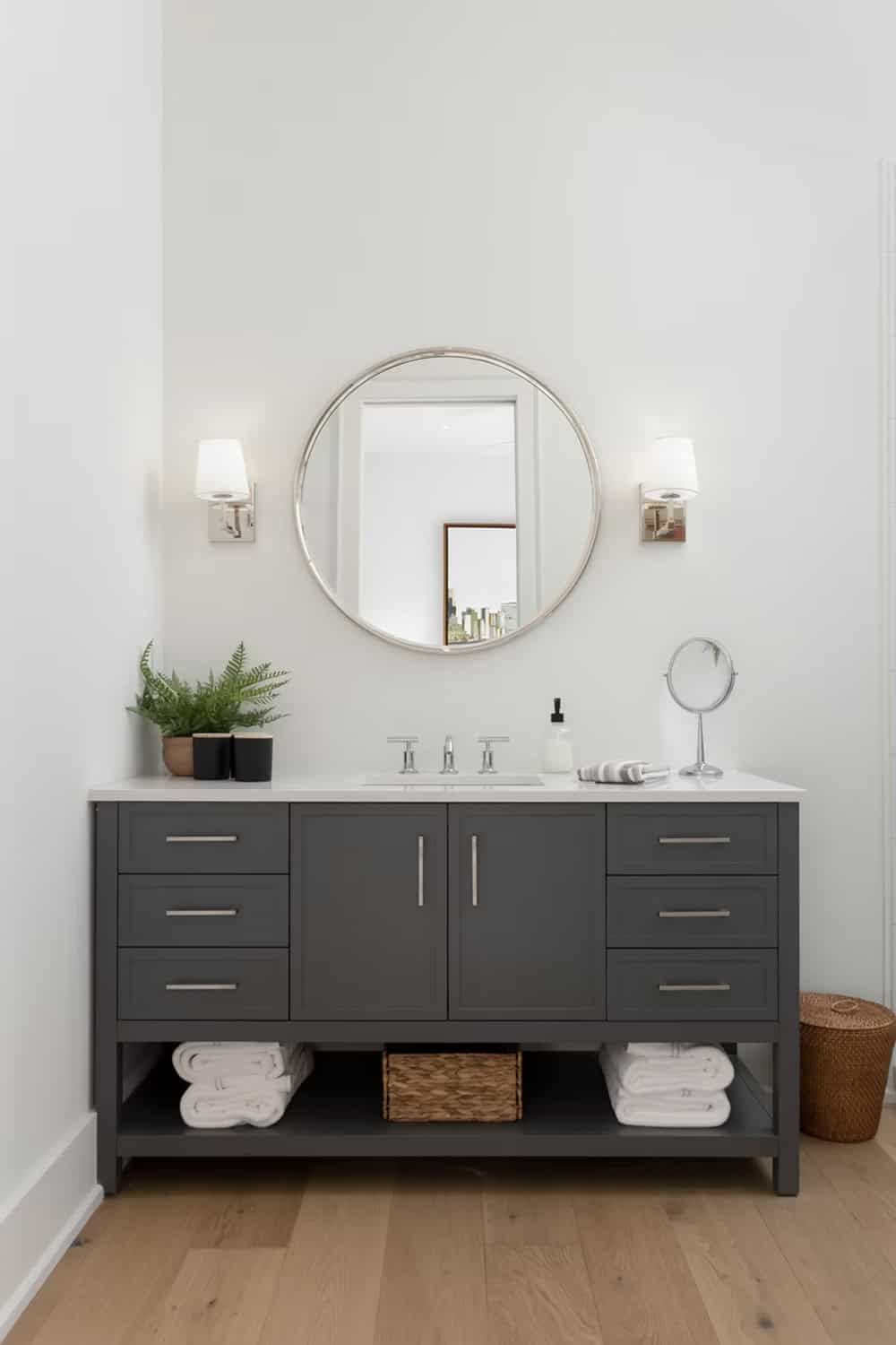 Close-up of dark bathroom vanity with round mirror, chrome faucet, wall sconces, and fern plant