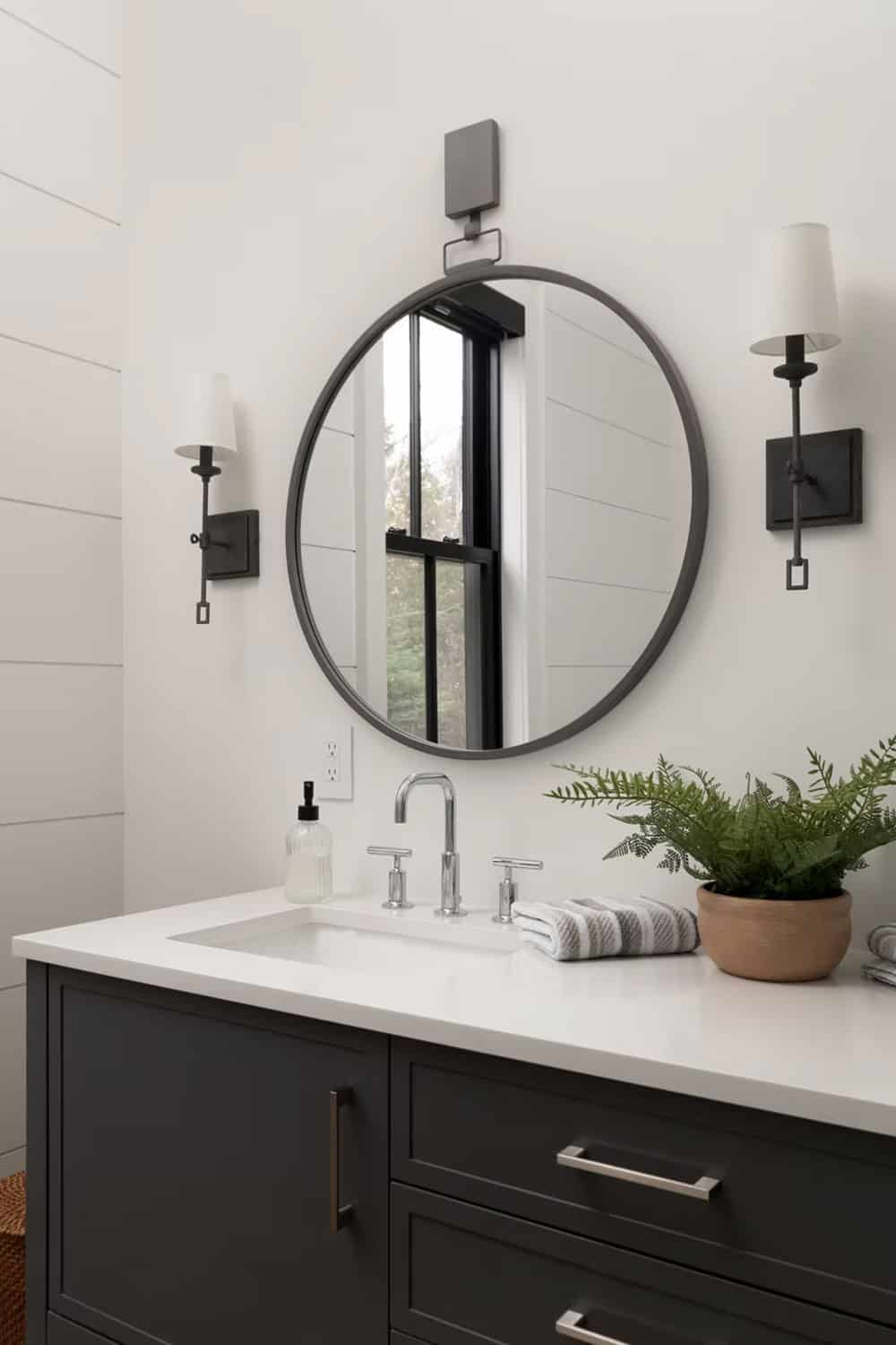 Close-up of dark bathroom vanity with large round mirror, black wall sconces, chrome faucet, and fern plant