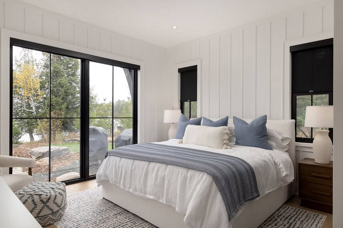 Guest bedroom with white board-and-batten walls, blue pillows, black-framed sliding glass doors to lakeside patio