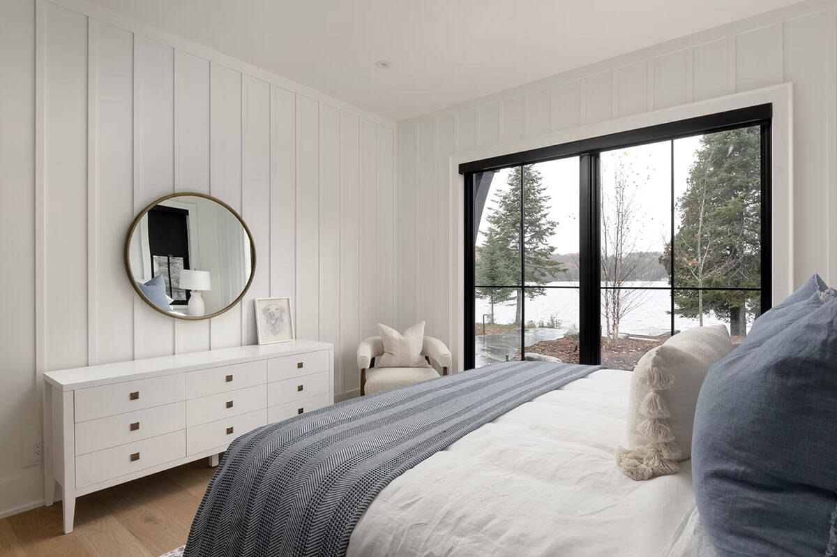Guest bedroom with white vertical shiplap, white dresser, round gold mirror, and black-framed lake view doors