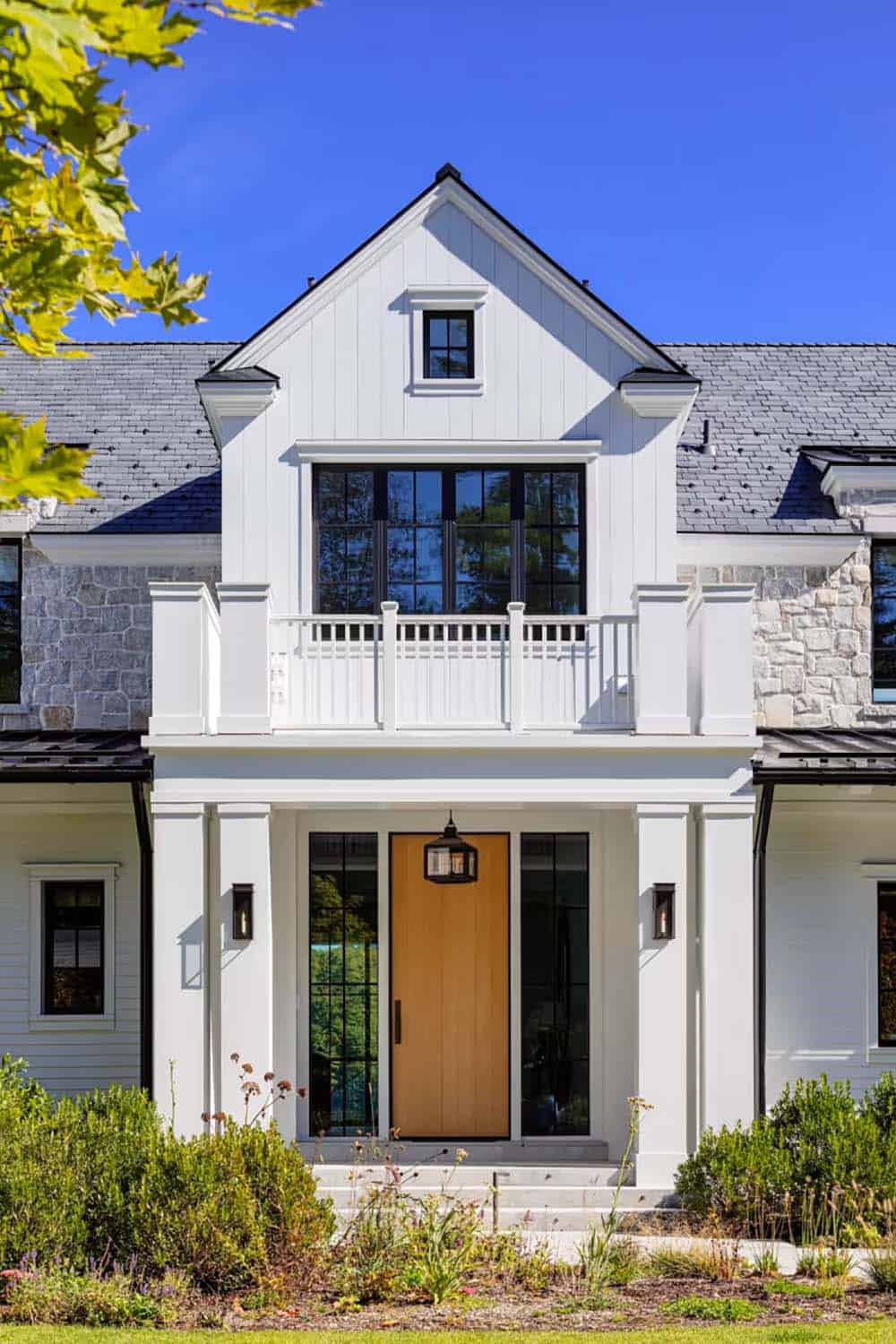 Front entrance of modern farmhouse with natural wood door, black-framed windows, white vertical siding, and upper balcony