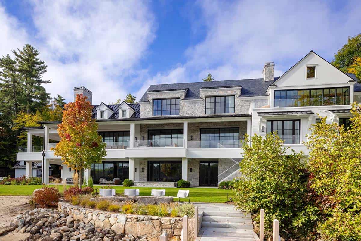 Rear facade of luxury lake house with stone and white exterior, large black-framed windows, terraced landscaping leading to private beach