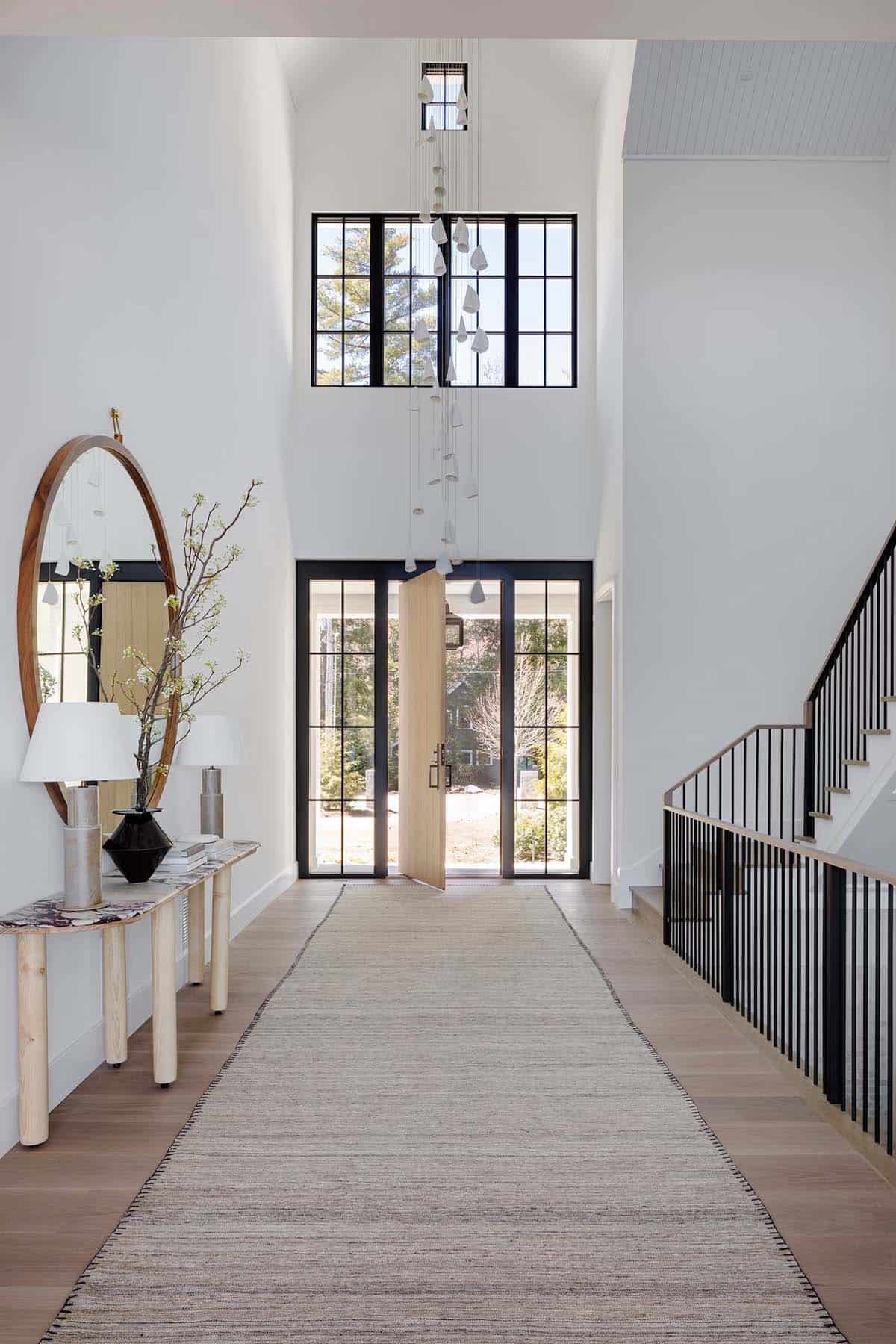 Double-height foyer with sculptural pendant chandelier, black-framed entry doors, light wood floors, and staircase with black iron railing