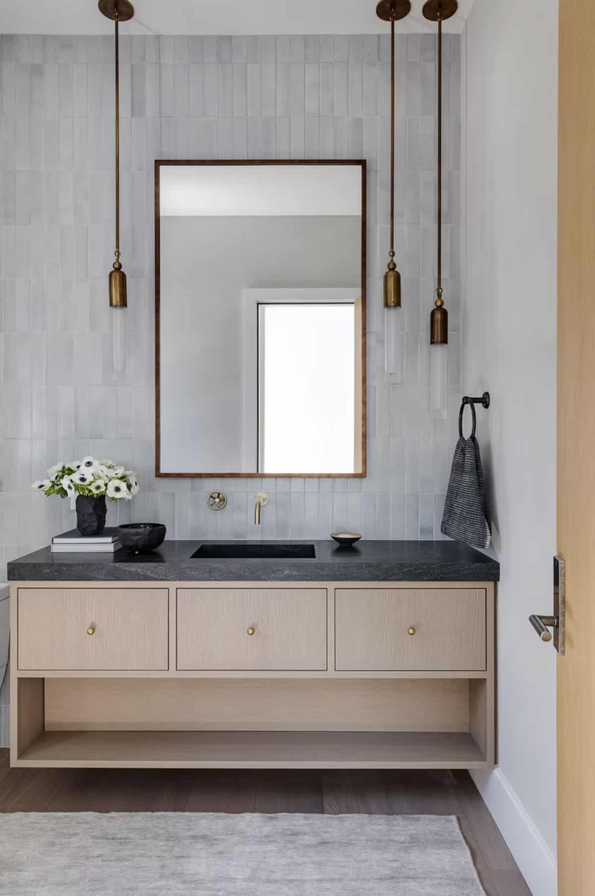 Modern powder room with gray zellige tile wall, floating oak vanity, dark stone countertop, and brass pendant lights