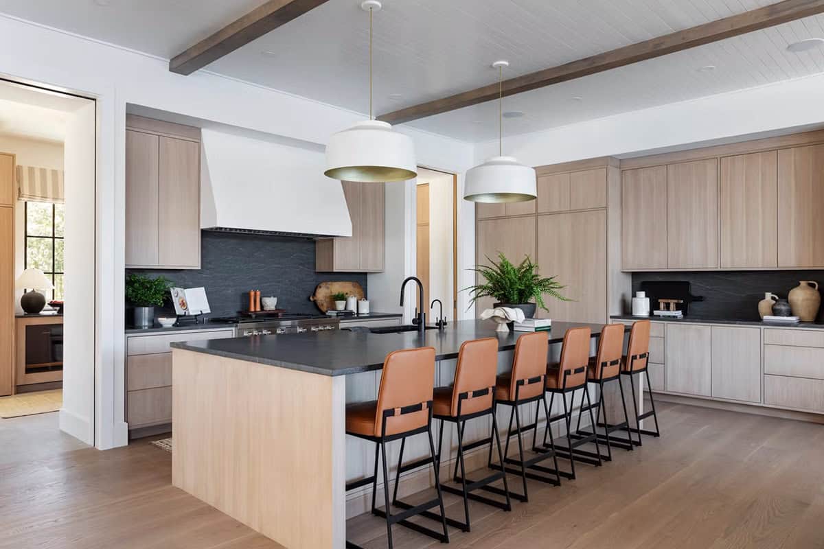 Open-concept kitchen with light oak cabinetry, dark soapstone countertops, leather bar stools, and white drum pendants over large island