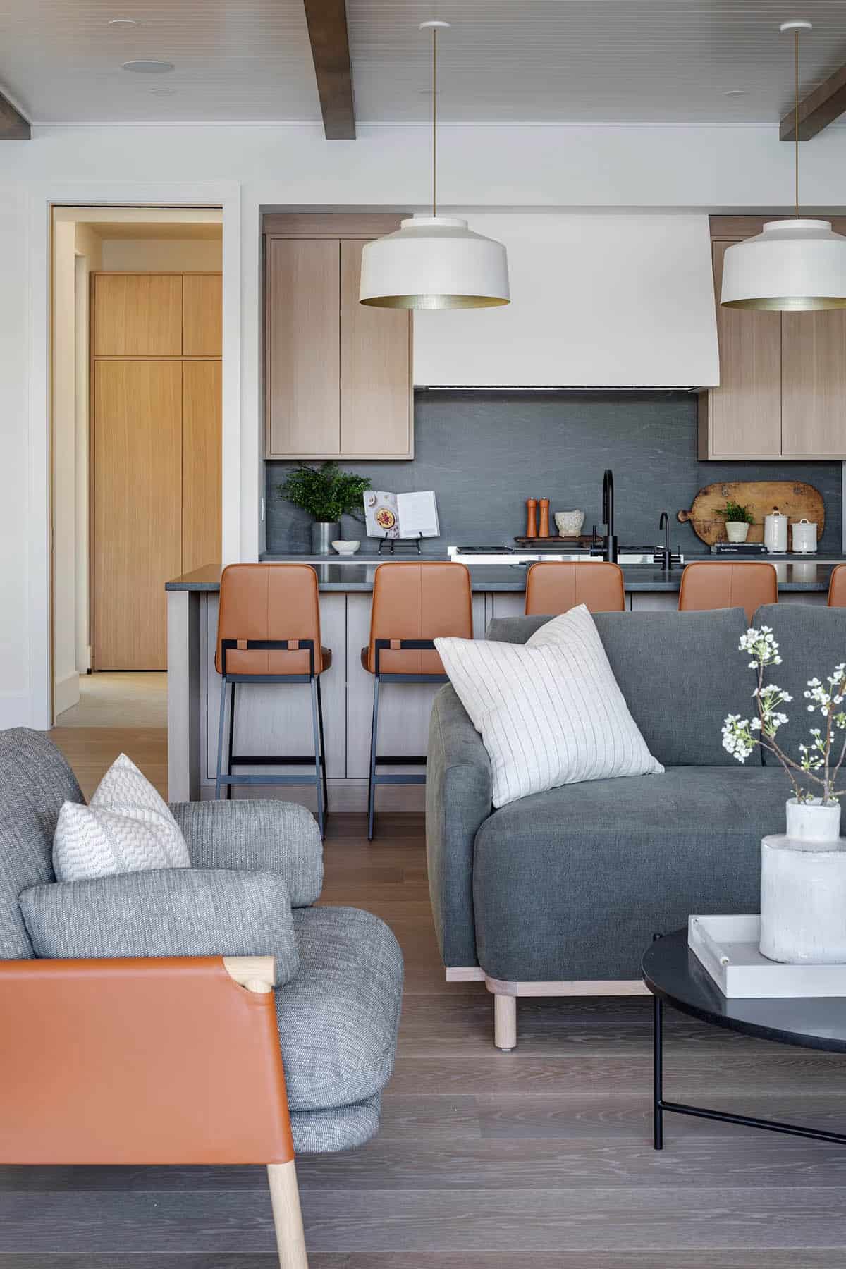 Kitchen and living area open plan with gray upholstered sofa, cognac leather chairs, oak cabinetry, and dark stone backsplash