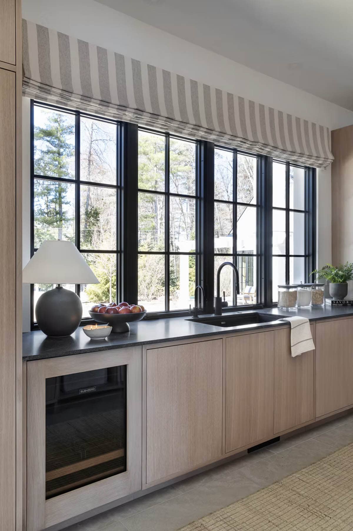 Kitchen sink area with black-framed grid windows, dark stone countertop, light oak cabinets, and striped Roman shade