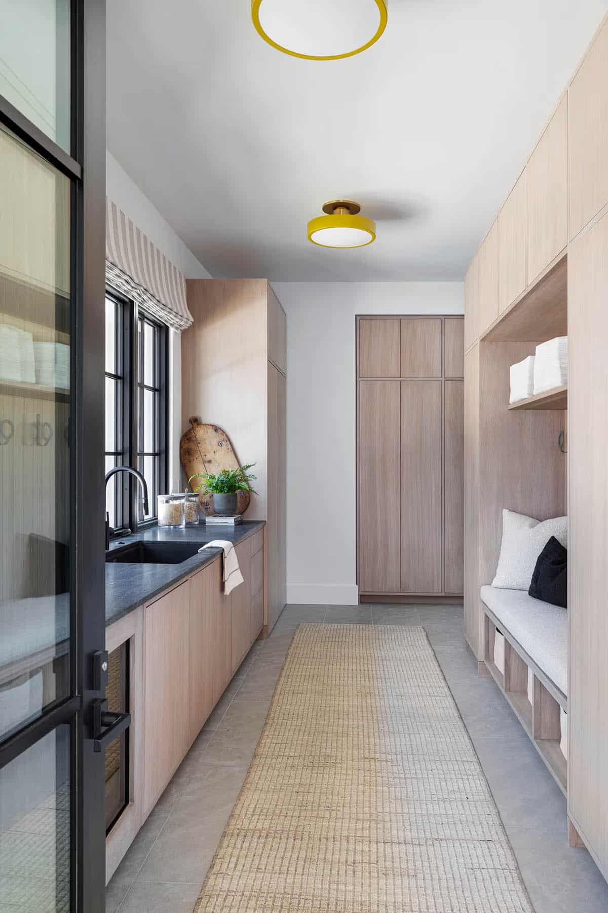 Mudroom with light oak built-ins, bench seating, yellow flush-mount ceiling lights, and black sink with dark stone counter
