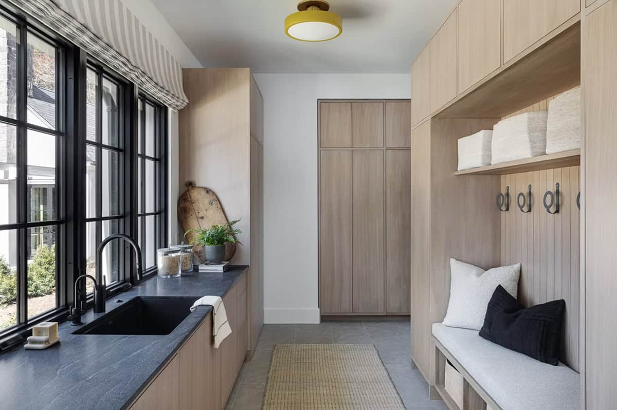 Bright mudroom with built-in coat hooks, upholstered bench, light oak storage, and black-framed windows with striped shade