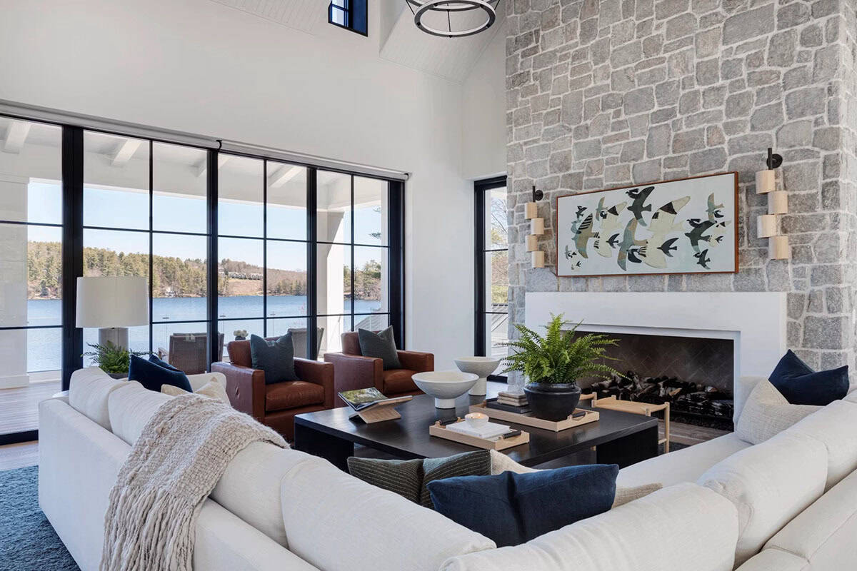 Living room with floor-to-ceiling lake views, white sectional sofa, cognac leather chairs, and full-height stone fireplace